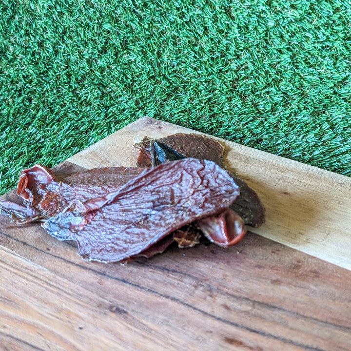A piece of lamb jerky placed on a wooden surface with a green background, illustrating the product's texture and color.